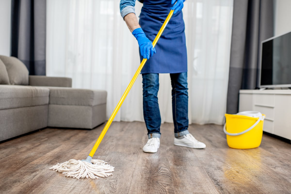 house cleaner wearing apron and rubber gloves mopping the wood floor of a livingroom
